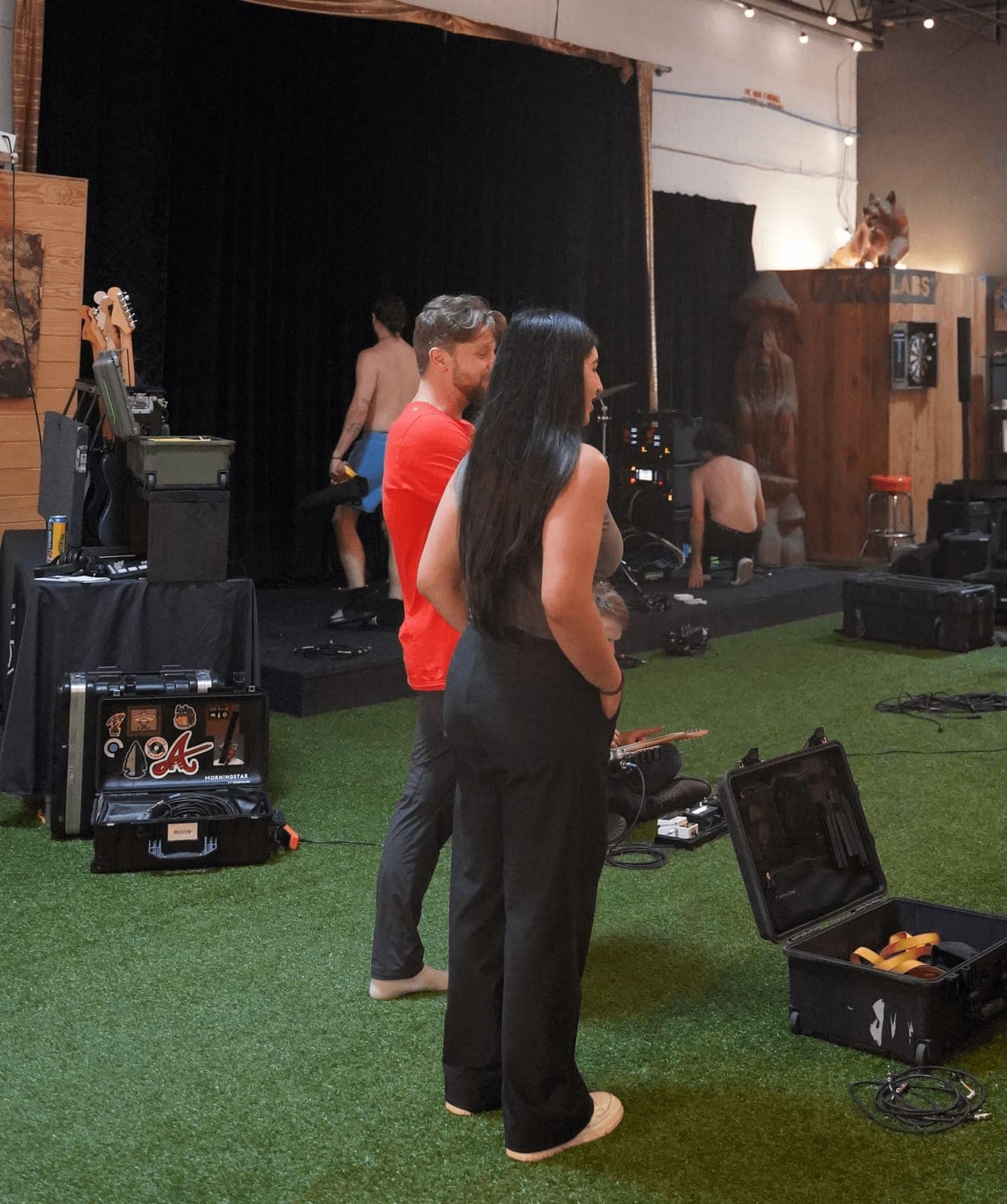 People set up gear on a green turf floor in front of a curtained stage before a show