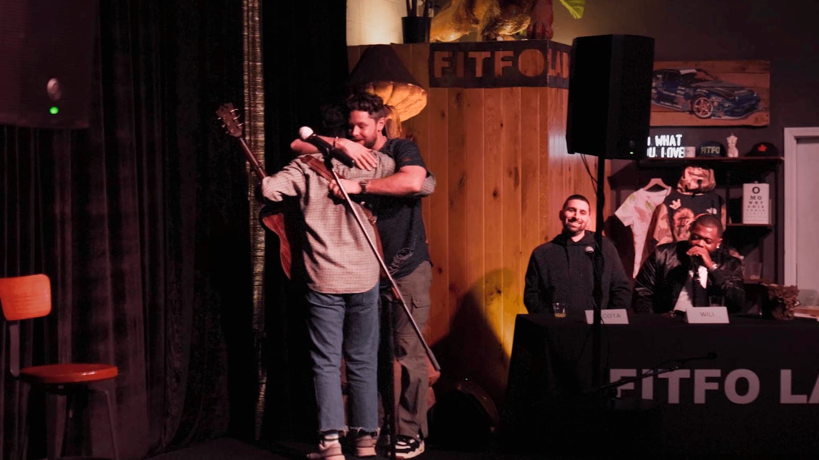 Two performers embrace on stage beside the judges' table at a Fitfo Labs showcase