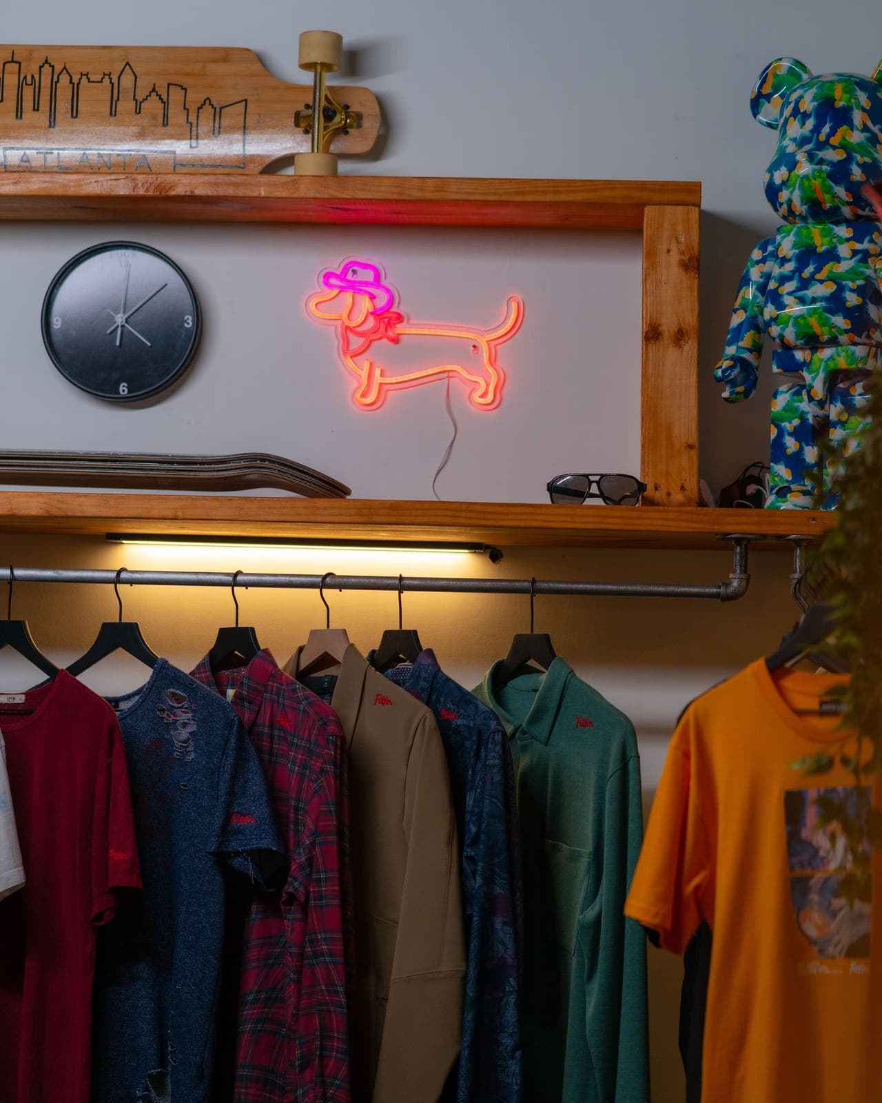 Shirts hang on a rack under a neon dog sign and a wall clock